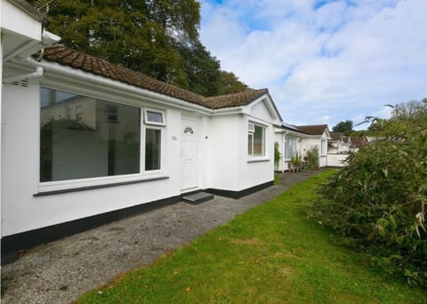 White bungalow with large front window, gravel driveway, and manicured lawn surrounded by trees and hedges