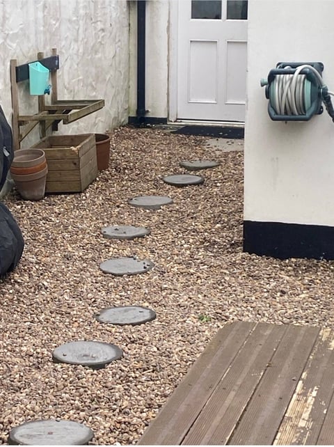 Gravel pathway with stepping stones leading to white door, flanked by storage items and a hose reel on the wall