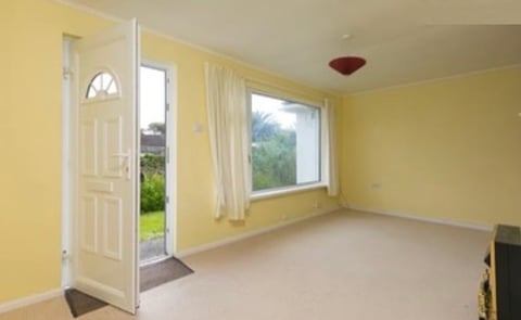 Bright yellow empty room with beige carpet, white doors opening to garden, large window with light curtains, and red ceiling fixture