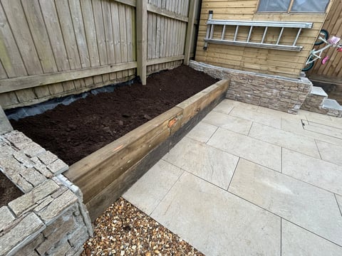 Wooden raised garden bed filled with dark soil in a paved backyard garden area with wooden fencing