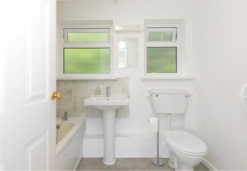 Clean, bright bathroom with white walls, pedestal sink, toilet, bathtub with tiling, and two windows with green frosted panes