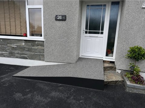 Modern residential entrance with white door, gray textured wall, black ramp, and small potted plants beside the doorway