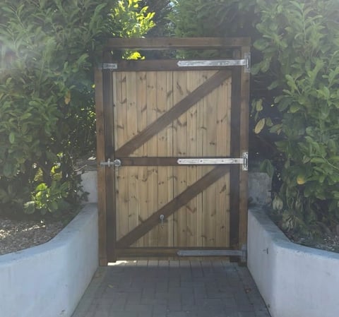 Wooden gate with diagonal braces in narrow passageway flanked by white walls and green plants