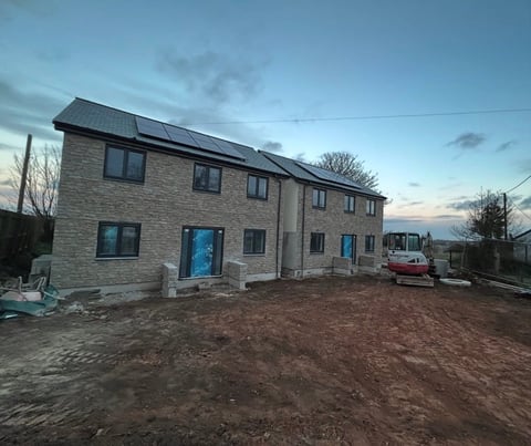 Modern stone house under construction with solar panels on roof, dirt driveway, and bare trees at dusk