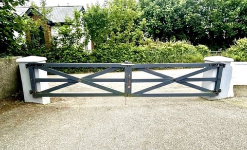 Black and white wooden farm gate with cross-brace design on a gravel driveway, surrounded by green hedges and trees
