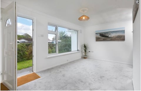 Empty bright room with white walls, light gray flooring, large windows overlooking gardens, and wooden door leading outside