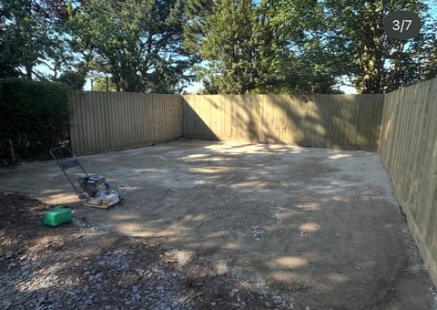 Empty residential backyard with concrete patio, metal fencing, scattered shade from trees, and gardening equipment