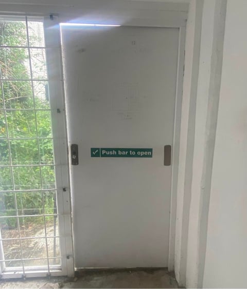 White push bar emergency exit door with green Push bar to open sign and window on left showing outdoor greenery