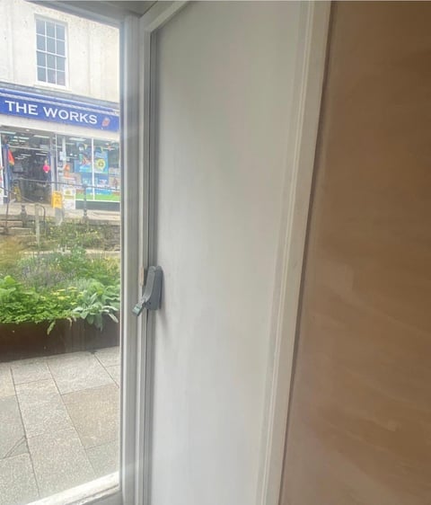 Open glass door with metal handle showing outdoor street view with The Works store sign and planters visible outside