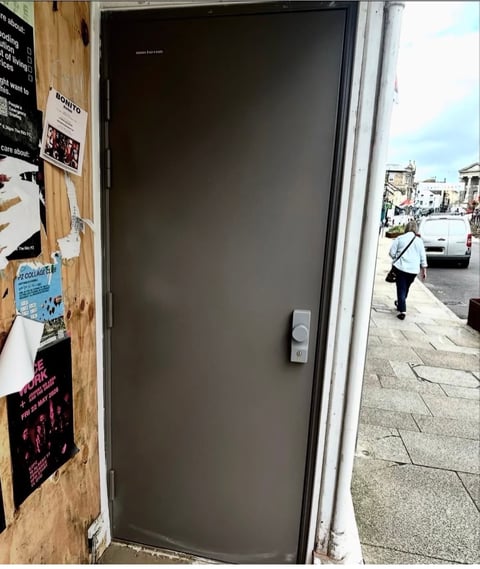Dark glass storefront door with white frame, posters on left wooden wall, and street view with pedestrian visible through windows