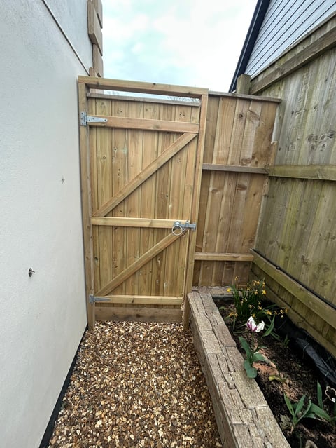 Wooden gate with diagonal bracing in narrow side yard with gravel ground and planter box with plants