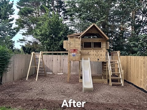 Wooden childrens playground structure with climbing ladders, slide, and treehouse-style cabin in fenced yard surrounded by trees