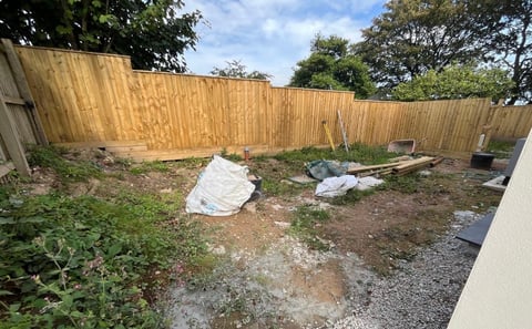 Overgrown backyard with wooden fence, scattered white tarps, gravel, dirt, and green vegetation under blue sky