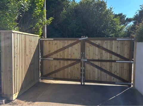 Wooden driveway gate with X-pattern design open on a concrete base, flanked by matching wooden fence panels with tree-lined background
