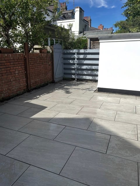 Paved backyard patio with gray stone tiles, brick walls, metal gate, and Victorian townhouse visible behind