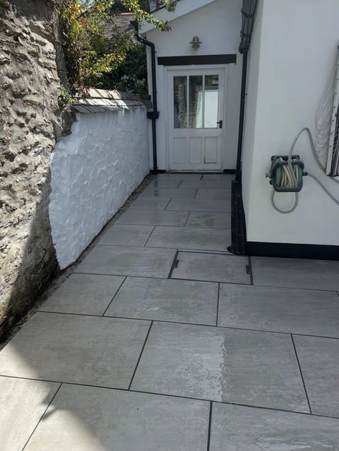 Narrow modern patio with gray slate tiles, white door, stone wall on left, and coiled hose on white wall