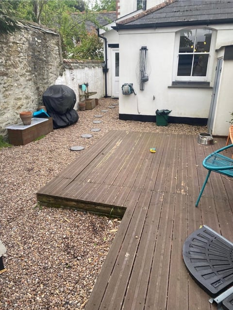 Residential backyard patio with wooden deck, gravel area, white cottage, garden items, and metal chair