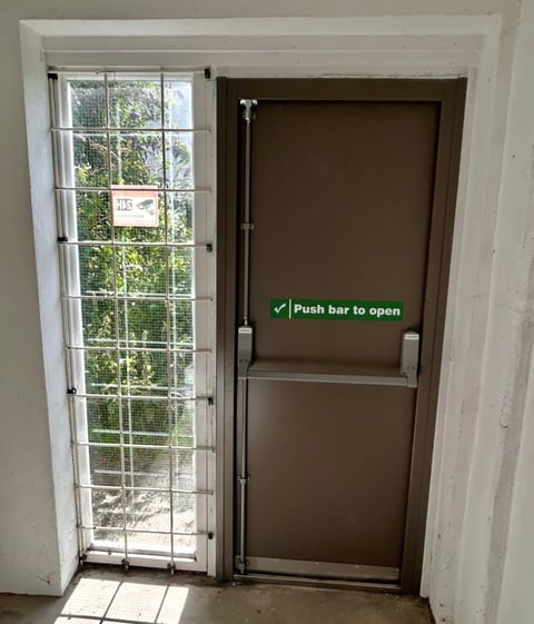 Emergency exit door with push bar mechanism next to a large window showing outdoor greenery and sunlight