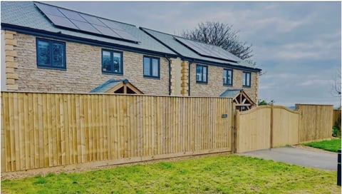 Modern stone house with blue-framed windows, slate roof, and tall wooden fence with driveway entrance