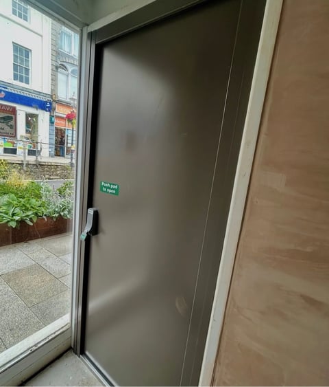 Gray push door with green sign inside storefront entryway, street view visible through adjacent window