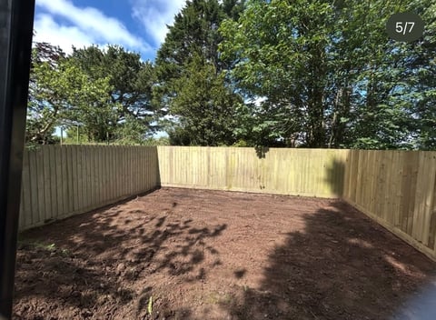 Empty backyard with wooden fence, brown mulch ground, and trees visible against blue sky