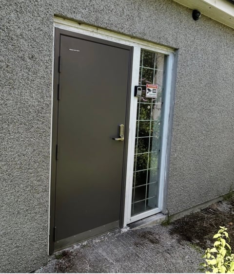 Black metal door with silver frame and small window beside it on a concrete wall