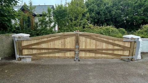 Wooden double-gate driveway entrance with white concrete pillars and arched cross-bracing design