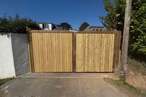 Wooden driveway gates with vertical slats, open entrance to residential property with house and vegetation visible in background