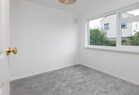 Empty bedroom with gray carpet, white walls, brass doorknob, ceiling light fixture, and window overlooking residential building