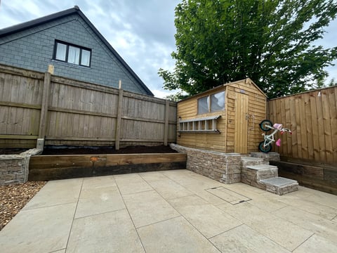 Backyard patio with paved stone flooring, wooden fencing, raised garden bed, and modern timber-clad garden building