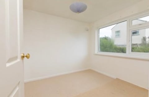 Empty bright bedroom with light wooden floor, white walls, brass door handle, ceiling light fixture, and large window overlooking greenery