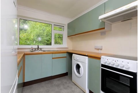 Modern kitchen with sage green cabinets, wooden countertops, white appliances, and large windows overlooking greenery